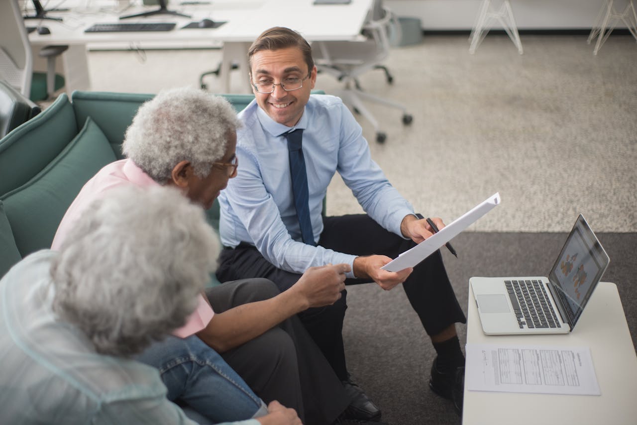 Home Consultant discussing financial plans with senior clients in a modern office setting, using documents and a laptop.